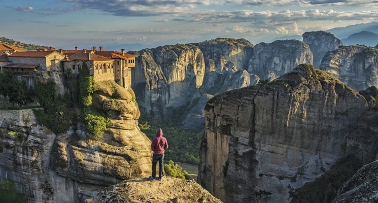 man standing on rocky cliff overlooking a monastery in Meteora Greece